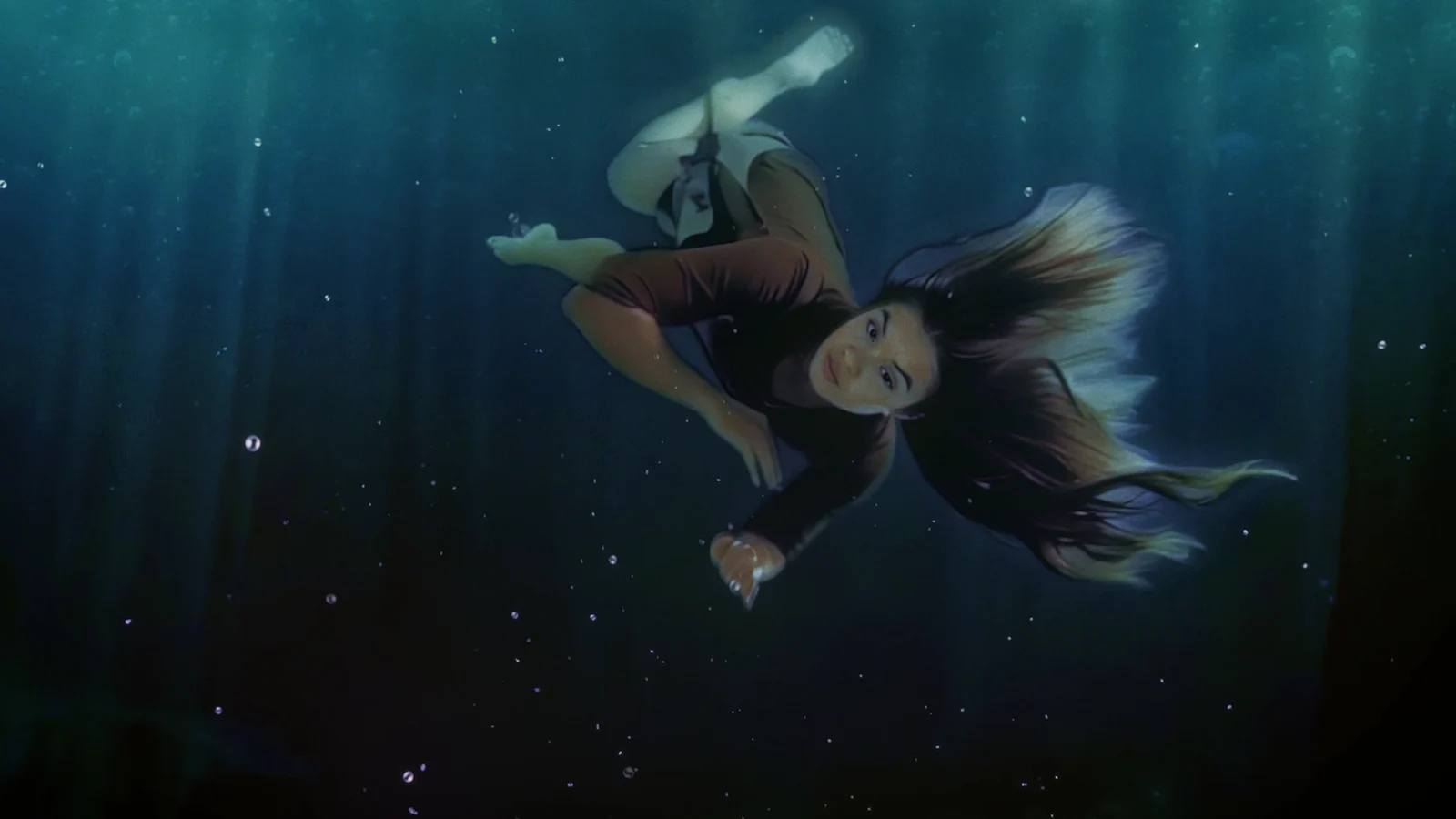 The woman turns underwater, her hair and clothing floating beneath shafts of blue light